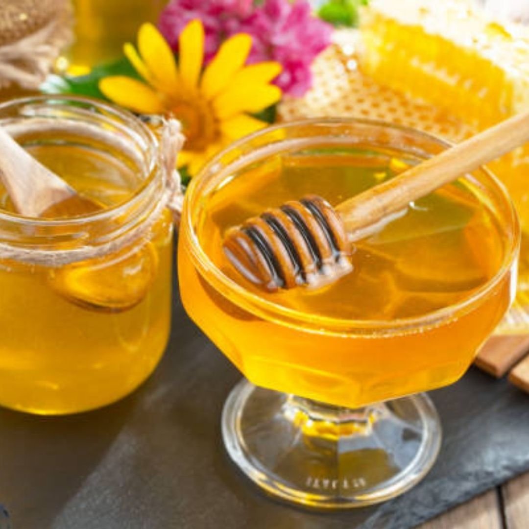 Close-up of golden honey in glass jar with honey dipper, fresh flowers in background — evokes natural, nostalgic essence from Elesaar’s first blog.